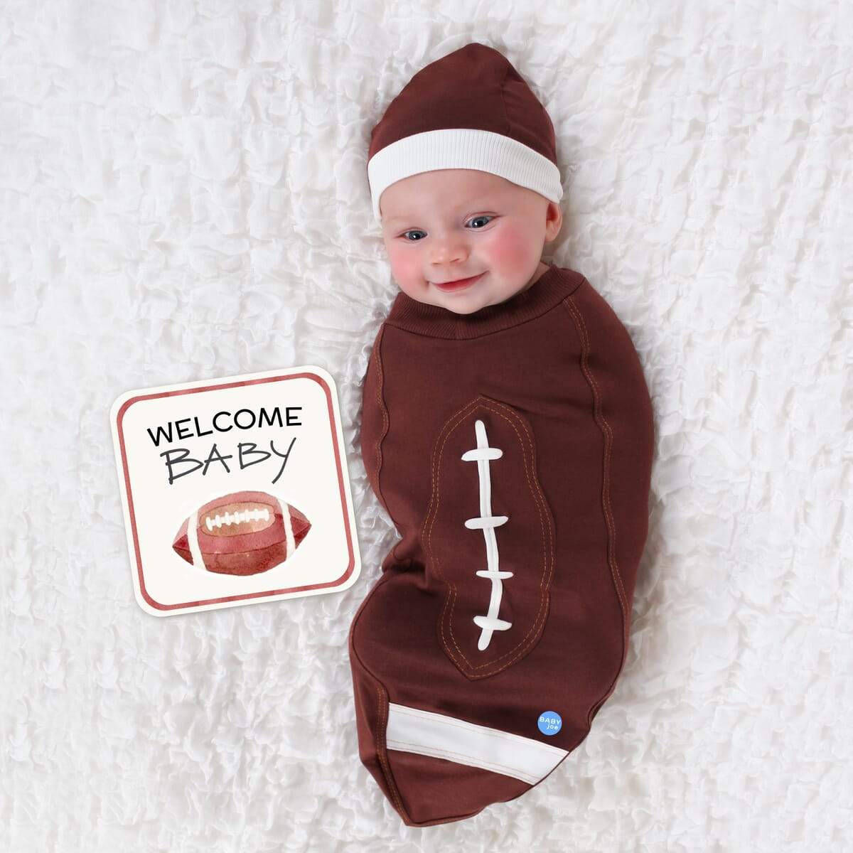 Newborn baby wrapped in football-themed baby cocoon swaddle with matching headpiece and announcement card on white textured blanket.