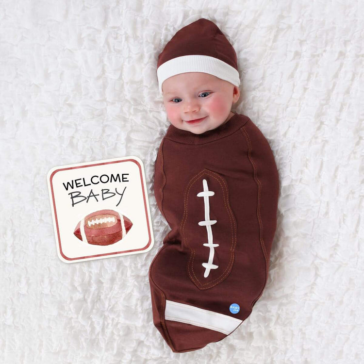 Newborn baby wrapped in football-themed baby cocoon swaddle with matching headpiece and announcement card on white textured blanket.